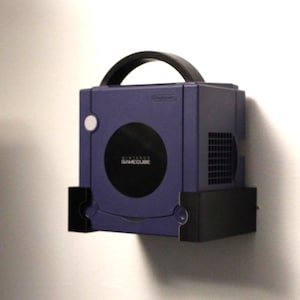 Puede incluir: Una consola Nintendo GameCube morada montada en la pared. La consola tiene un asa negra y una puerta circular para la unidad de disco. El GameCube está asegurado por un soporte de pared negro.