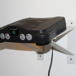 May include: A black Nintendo 64 console mounted on a white wall shelf. The console features the Nintendo 64 logo and four controller ports. The shelf is fixed to the wall with screws, and a black power cable is visible.
