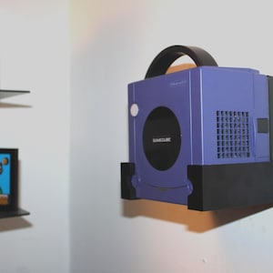 May include: A purple Nintendo GameCube console mounted on a wall. The console has a black handle and a black base. The GameCube logo is visible on the front. A shelf with a video game is visible in the background.