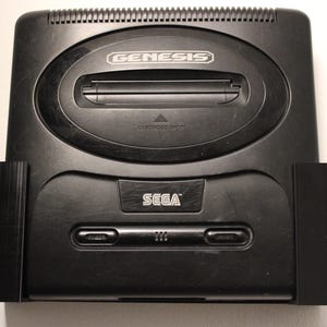 以下が含まれることがあります： 黒いセガジェネシスコンソール。「GENESIS」の文字がカートリッジスロットの上に銀色の文字で表示されています。「SEGA」の文字は、電源ボタンとリセットボタンの下に銀色の文字で印刷されています。コンソールは長方形で、上部が湾曲しています。