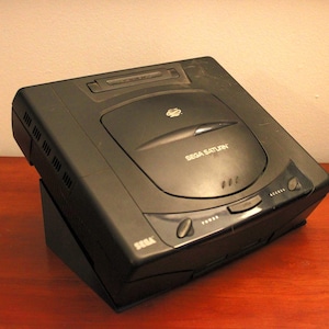 May include: A black Sega Saturn video game console. The console is rectangular with rounded edges and features the Sega Saturn logo. The console has a disc tray, power and access buttons, and the words "Sega Saturn" are printed on the front.