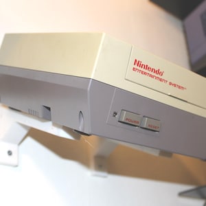 May include: A vintage Nintendo Entertainment System console in gray and cream colors. The console has the Nintendo logo and the words "ENTERTAINMENT SYSTEM" in red. It features power and reset buttons.