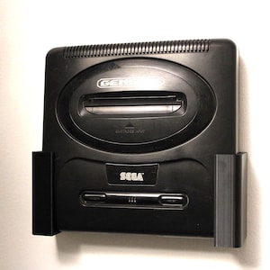 May include: A black Sega Genesis console mounted on a white wall. The console has the Sega logo and the word "GENESIS" printed on it. The console is a classic video game system.