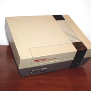 May include: A vintage Nintendo Entertainment System console in beige and gray. The console has a rectangular shape with a slot for game cartridges and the text "Nintendo ENTERTAINMENT SYSTEM" in red. The console is on a wooden surface.