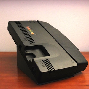 May include: Black TurboGrafx-16 video game console, angled on a black stand. The console has the TurboGrafx-16 logo in red, yellow, and green. The console is on a wooden surface.