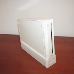 Może przedstawiać: Biała konsola do gier Nintendo Wii, stojąca pionowo na pasującym stojaku. Konsola jest prostokątna z gniazdem na płyty z grami i logo Wii z przodu. Konsola stoi na ciemnobrązowej drewnianej powierzchni.