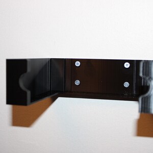 May include: A black, L-shaped wall-mounted shelf with rounded cutouts on the ends. The shelf is attached to the wall with four silver screws. The shelf is made of a dark material and is mounted on a white wall.