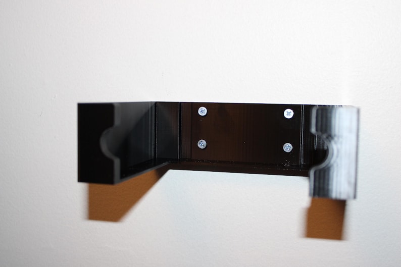 May include: A black, L-shaped wall-mounted shelf with rounded cutouts on the ends. The shelf is attached to the wall with four silver screws. The shelf is made of a dark material and is mounted on a white wall.