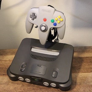 Op de afbeelding: Een grijze Nintendo 64-console met een bijpassende controller. De controller heeft een D-pad, rode, groene, blauwe en gele knoppen en een centrale analoge stick. De console is donkergrijs en staat op een houten oppervlak.