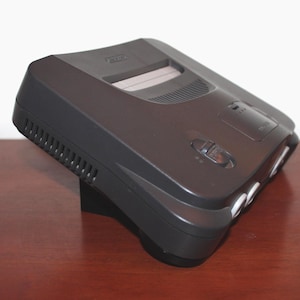 May include: A dark gray Nintendo 64 video game console. The console has a rectangular shape with rounded edges and several buttons and ports. The top of the console has a slot for game cartridges. The console is sitting on a brown surface.