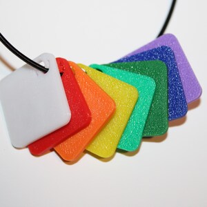 May include: A colorful necklace featuring a series of square pendants in a rainbow of colors: white, red, orange, yellow, green, blue, and purple. The pendants are attached to a black cord, creating a vibrant and eye-catching accessory.