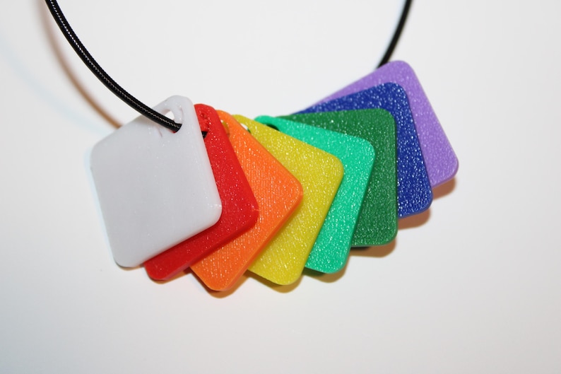 May include: A colorful necklace featuring a series of square pendants in a rainbow of colors: white, red, orange, yellow, green, blue, and purple. The pendants are attached to a black cord, creating a vibrant and eye-catching accessory.