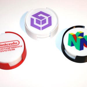 Puede incluir: Juego de tres posavasos redondos con logotipos de consolas de videojuegos clásicas. Uno tiene el logotipo de Nintendo Entertainment System en rojo, otro el logotipo púrpura de GameCube y el tercero el logotipo colorido de Nintendo 64.