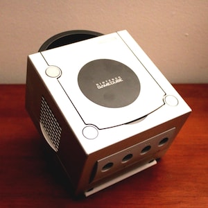 May include: Silver Nintendo GameCube console with a black disc tray and the GameCube logo. The console has a silver top with a black circle in the center. The front of the console has four controller ports.