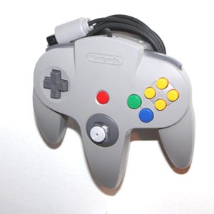 May include: A gray Nintendo 64 controller with a black cable. The controller features a directional pad, a central joystick, and colored buttons: red, green, blue, and yellow. The Nintendo logo is visible.
