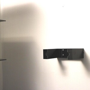 May include: A black wall-mounted shelf with a rectangular design. The shelf is attached to a white wall with screws. The shelf is designed to hold items. The image is taken in a room with a white wall.