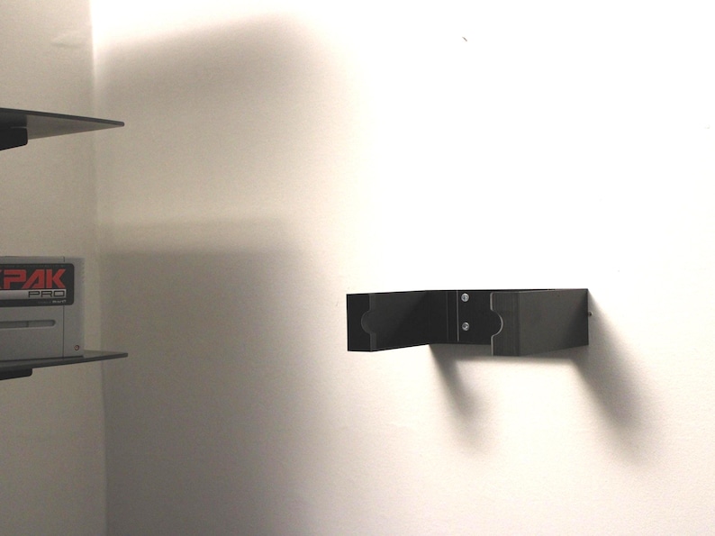 May include: A black wall-mounted shelf with a rectangular design. The shelf is attached to a white wall with screws. The shelf is designed to hold items. The image is taken in a room with a white wall.