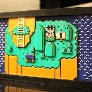 May include: A framed pixel art piece depicting a level from a classic video game. The artwork features a green landscape with a castle, platforms, and a number 1. The background includes blue and yellow sections, all within a black frame.