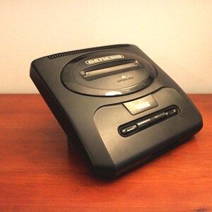 May include: A black Sega Genesis video game console. The console has a rectangular shape with a cartridge slot on top and a controller port on the front. The Sega logo is visible.