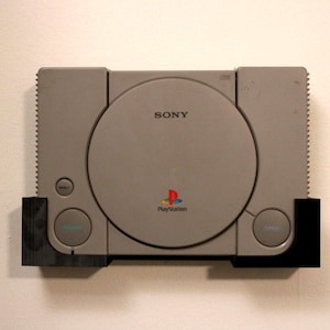 May include: A gray PlayStation console with the Sony logo and PlayStation logo. The console has a disc tray and several buttons, including power and reset. The console is mounted on a black bracket.
