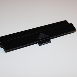 Puede incluir: Una cubierta negra rectangular de plástico para el compartimento de la batería con una pequeña pestaña y un diseño triangular. La cubierta está diseñada para adaptarse a un dispositivo electrónico específico.