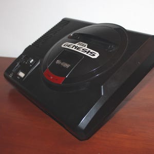May include: Black Sega Genesis 16-bit video game console. The console has a rounded top with the Sega Genesis logo and a red accent. The console is on a brown surface.