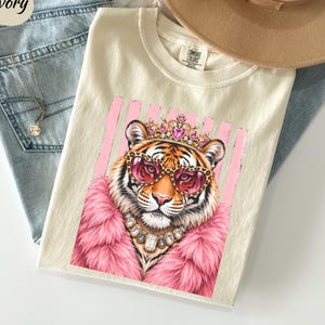 May include: An ivory t-shirt featuring a graphic of a tiger wearing a pink fur coat, a jeweled crown, leopard print heart-shaped sunglasses, and a crystal necklace. The design is set against a background of vertical pink stripes on the fabric.