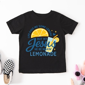 A black t-shirt with a colorful graphic design, including a lemon slice, a drink, and text that says "Life gave me some lemons but my Jesus he be making lemonade".