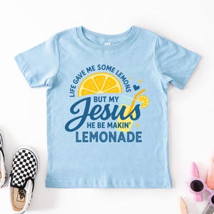 A light blue t-shirt with a graphic design and text, along with a pair of black and white checkered shoes, a pair of sunglasses, and some other objects on a white background.