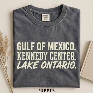 May include: A charcoal grey short-sleeve t-shirt with a crew neckline. The shirt features the white text GULF OF MEXICO. KENNEDY CENTER. LAKE ONTARIO. printed on the front. The interior neck label reads COMFORT COLORS and includes size S-P-CH.