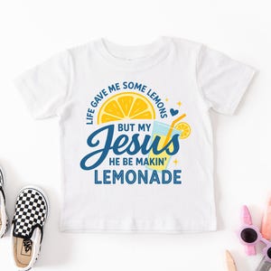 A white t-shirt with a colorful graphic design, including a lemon slice, a cup, and text that says "Life gave me some lemons, but my Jesus he be making lemonade".