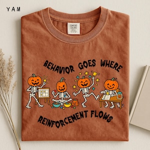 May include: A rust-colored t-shirt featuring a graphic of four skeletons with pumpkin heads. The text reads BEHAVIOR GOES WHERE REINFORCEMENT FLOWS. The skeletons are shown in various activities including using a whiteboard, sitting with bowls, dancing, and writing at a desk with a jar.