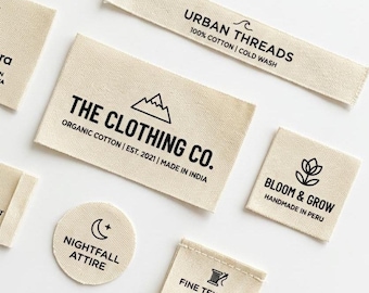 May include: Assortment of beige fabric clothing labels with black text. Labels include "URBAN THREADS", "THE CLOTHING CO.", "BLOOM & GROW", "NIGHTFALL ATTIRE", and "FINE TEXTILES". Some labels feature mountain, floral, and moon graphics.