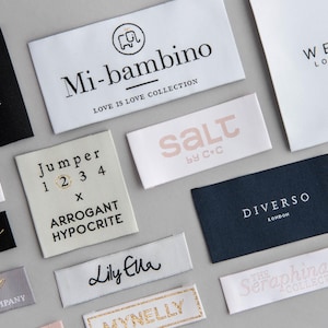 Op de afbeelding: Een verzameling stoffen labels met verschillende merknamen en logo's. De labels zijn in verschillende kleuren, waaronder zwart, wit en grijs, met tekst in goud, zwart en roze. Merken zijn onder meer "ANASTASIA LONDON", "Mi-bambino" en "WEAR LONDON".