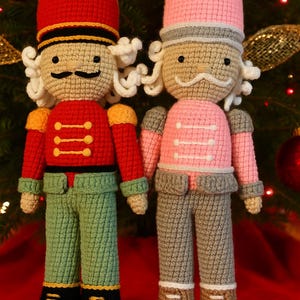 May include: Two handmade crochet nutcrackers. One wears a red uniform with gold accents and green trousers. The other wears a pink uniform with grey trousers. Both have white curly hair and moustaches. Christmas decor.