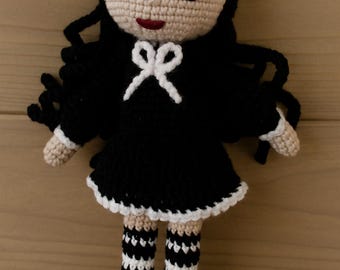 May include: A handmade crochet doll with black hair and a black dress. The doll has a cream-coloured face, arms, and legs, with black and white striped socks. A white bow adorns the dress. The doll is approximately 25 cm tall.