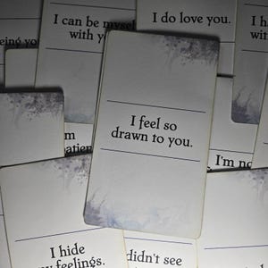 Peut inclure: Une collection de cartes rectangulaires blanches avec du texte noir et de subtiles touches d'aquarelle grise. La carte centrale affiche la phrase 'I feel so drawn to you.' D'autres cartes visibles portent des phrases comme 'I do love you,' 'I can be myself with you,' et 'I hide my feelings.'