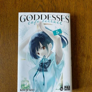 May include: A manga volume titled "Goddesses Cafe Terrace" with a cover illustration of a young woman with dark hair. The number "5" is visible on a tag. The author's name, Kouji Seo, is printed at the bottom.