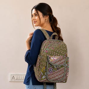 May include: A quilted backpack featuring an olive green base with a multi-colored floral and geometric pattern. The bag includes a front pocket with a flap, side pockets, and striped fabric trim along the edges, straps, and pocket seams.