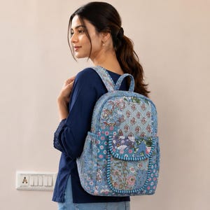 May include: A quilted backpack featuring a patchwork design in shades of blue, pink, and white with floral and paisley patterns. The bag includes a front pocket with a flap, a top handle, and two shoulder straps, all finished with blue piping.