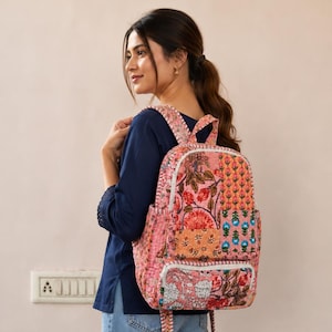 Puede incluir: Una mochila acolchada con un diseño de patchwork que presenta varios patrones florales y botánicos en tonos rosa, naranja y verde. La bolsa incluye un compartimento principal con cremallera, un bolsillo frontal más pequeño y dos correas con costuras blancas.