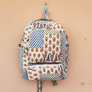 May include: A patchwork backpack with a variety of floral and geometric patterns in shades of white, blue, orange, and pink. The backpack is hanging on a peach-colored wall. It has a front pocket and adjustable straps.