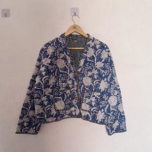 May include: A blue and white floral print jacket with wooden buttons and two front pockets. The jacket has a black and white striped lining and is hanging on a wooden hanger.