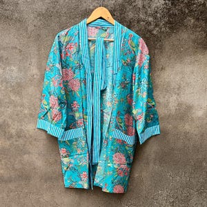 May include: A turquoise floral print robe with a striped trim and matching tie closure. The robe features a vibrant pattern of pink flowers and green leaves. The robe is hanging on a wooden hanger.
