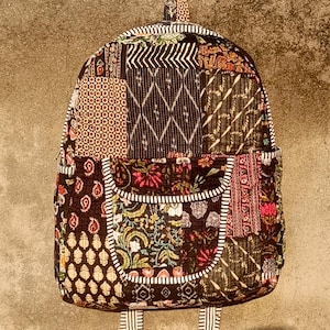 Puede incluir: Una mochila patchwork con una variedad de patrones en tonos marrones, negros y rojos. La mochila tiene dos bolsillos delanteros y correas a rayas. El asa superior también tiene un diseño. La mochila cuelga contra una pared neutra.