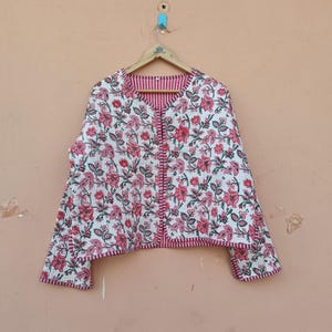 May include: A white floral jacket with pink and red flowers, hanging on a wooden hanger. The jacket has a quilted texture and a pink striped lining. The sleeves are three-quarter length.