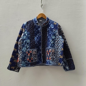 May include: A blue patchwork quilted jacket with floral and geometric patterns. The jacket has two pockets and a striped collar. The jacket is hanging on a wooden hanger. The jacket is in shades of blue.