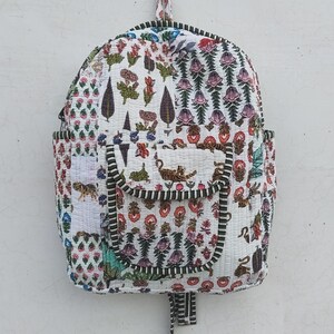 Puede incluir: Una mochila de tela acolchada con base blanca y un estampado floral y de tigres multicolor. La bolsa incluye un bolsillo frontal con solapa, un asa superior y ribetes a rayas de color verde oscuro y blanco en los bordes y costuras.