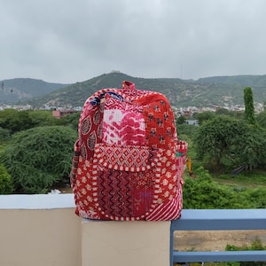 May include: A quilted backpack featuring a patchwork design in shades of red, pink, and white. The bag displays various intricate patterns including floral, geometric, and zigzag motifs, with a front pocket and a top handle for carrying.