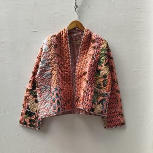 May include: A quilted jacket with a patchwork design. The jacket features a mix of floral and geometric patterns in shades of pink, peach, and brown. It has a relaxed fit and is hanging on a wooden hanger.
